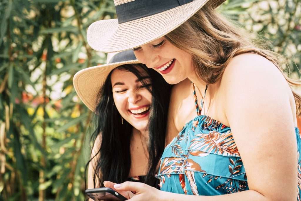 Two beautiful and cheerful female tourists in straw hat having fun text messaging using phone